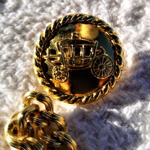 May include: Close-up of several gold buttons featuring a carriage design. The buttons have a rope-like border and are set against a white, textured background. A gold chain is also visible.