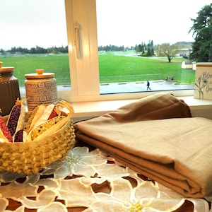 May include: A still-life arrangement featuring a woven basket filled with colorful dried corn, two ceramic jars with wooden lids, and a folded tan blanket. A small decorative box with the text "Bloom and Grow" is also present.