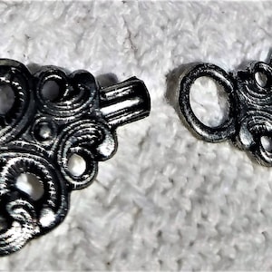 May include: Two-part silver-coloured clasp with an ornate, swirling design. The clasp has a hook and eye closure, with a detailed, openwork pattern. The metal has a slightly aged appearance, suggesting a vintage or antique style. Suitable for jewellery making or craft projects.