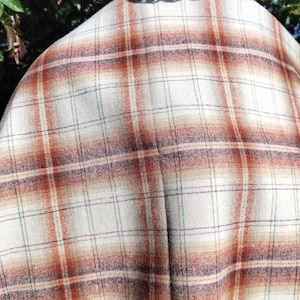 May include: Close-up of a plaid fabric in cream, rust, and blue. The pattern features a grid of intersecting lines in rust and blue over a cream background. The fabric appears to be a woven material, possibly wool or a wool blend.
