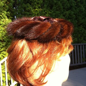 May include: A vintage brown fur hat with a dark brown satin bow. The hat is circular and sits atop a head with auburn hair. The background is a green hedge and a wooden deck.