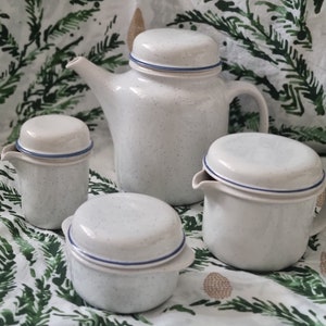 May include: A white ceramic tea set with blue trim. The set includes a teapot, creamer, sugar bowl, and lid. The set is on a white background with a green floral pattern.