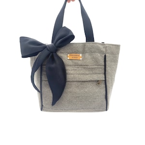 Può includere: Una borsa tote grigia con un grande fiocco nero sul davanti. La borsa ha manici neri e una piccola tasca con cerniera. Una piccola etichetta rettangolare è attaccata alla borsa.