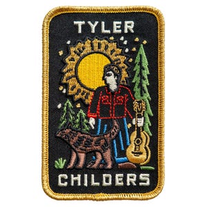 Puede incluir: Un parche bordado rectangular con fondo negro y borde dorado. Muestra a una persona con camisa roja y pantalones azules de pie junto a un perro y una guitarra. Un sol amarillo y árboles verdes aparecen al fondo. El texto TYLER CHILDERS está bordado en blanco.