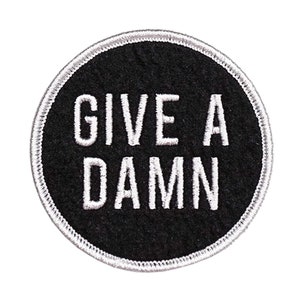 Op de afbeelding: Ronde zwarte patch met de witte tekst "GIVE A DAMN" in vette letters. De patch heeft een wit gestikte rand en staat op een witte achtergrond. Een draagbaar accessoire.