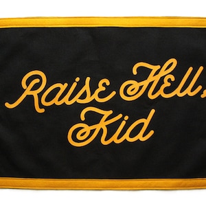 May include: Black canvas banner with a gold border and the text "Raise Hell, Kid" in a gold cursive font.