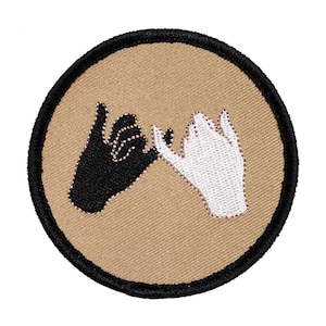 May include: A round, tan fabric patch with black stitching around the edges. Two hands, one black and one white, are embroidered on the patch, with their pinky fingers intertwined.