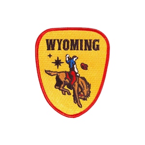 Op de afbeelding: Gele en rode geborduurde patch met het woord "WYOMING" in zwarte letters. In het midden van de patch is een cowboy te zien die op een bokkend paard rijdt.