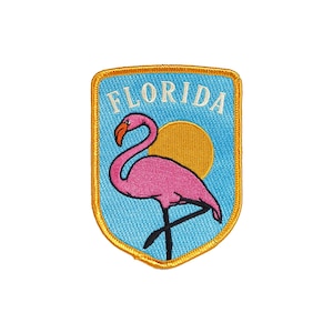 Könnte beinhalten: Ein blauer und goldener gestickter Aufnäher mit einem rosafarbenen Flamingo und einer gelben Sonne. Der Text "FLORIDA" ist in Weiß auf den Aufnäher gestickt.