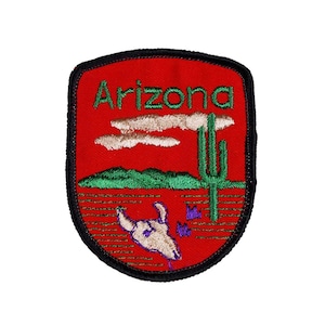 May include: Red embroidered patch with the word "Arizona" in white lettering. The patch features a green cactus, a brown mountain range, and a white cow skull with purple flowers.