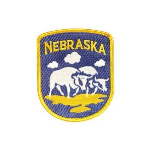 May include: A blue and yellow embroidered patch with the word "NEBRASKA" in yellow lettering. The patch features a white silhouette of three cows in a field.
