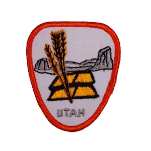 May include: A white embroidered patch with an orange border. The patch features a stylized image of a mountain range, wheat stalks, and a beehive. The text "UTAH" is embroidered in the bottom center of the patch.