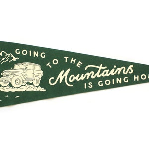 以下が含まれることがあります： 白い文字で「Going to the Mountains is going home」と書かれた緑色のペンナント旗。旗の左側には、山道を走るジープの白いイラストが描かれている。