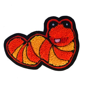 May include: A colorful, embroidered patch in the shape of a cartoon worm. The worm features a textured, orange body with yellow and orange stripes, black eyes, and a pink tongue. The patch is outlined in black.