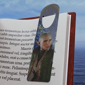 Puede incluir: Un abridor de botellas de metal con forma de marcapáginas que presenta un personaje de fantasía con el pelo corto y oscuro y un atuendo verde y marrón. El personaje se representa sobre un fondo de escena de bosque con un cielo azul y nubes blancas.