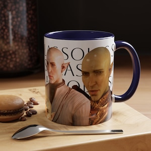 May include: A white ceramic mug with a blue rim and handle. The mug features a graphic of two bald, elf-like characters with the text "Lassols as Acolytes" printed on the mug.