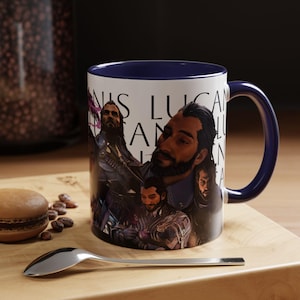 May include: White ceramic mug with a navy blue interior and handle. The mug features a collage of images of a man with a beard and the text "NIS LUCAN". A spoon and a macaron are on a wooden surface.