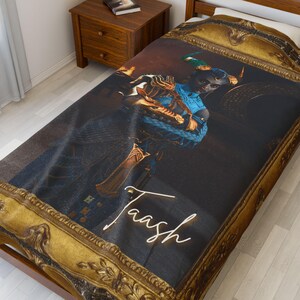 May include: A dark blue and gold throw blanket with a framed image of a person with horns and a sword. The image is set against a dark background and the person's name, "Taash", is written in white text below the image.