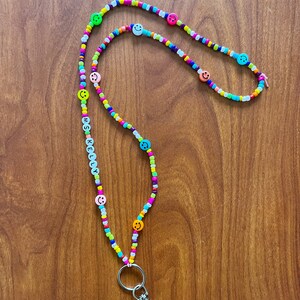 May include: A colorful beaded lanyard with the name "MS. KELLY" spelled out in white letter beads. The lanyard features a variety of small, brightly colored beads and smiley face beads. A silver-colored clasp and key ring are attached at the bottom.