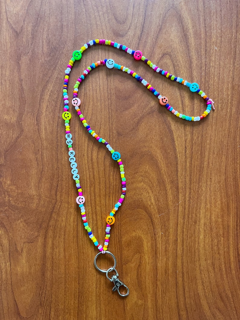 May include: A colorful beaded lanyard with the name "MS. KELLY" spelled out in white letter beads. The lanyard features a variety of small, brightly colored beads and smiley face beads. A silver-colored clasp and key ring are attached at the bottom.