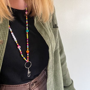 May include: A beaded lanyard with the name "KELLY" spelled out in white beads. The lanyard features colorful beads, smiley face beads, and a silver clasp. The lanyard is worn over a black shirt and a green corduroy jacket.