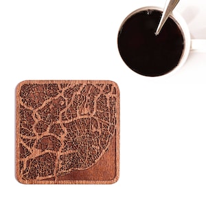 May include: A square, wooden coaster with a detailed map design etched into its surface. The coaster is a warm brown color and has rounded corners. A white mug with dark liquid and a spoon is in the upper right corner.