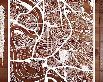 Puede incluir: Un recorte de papel blanco de un mapa de la ciudad con un río serpenteando a través de él. El mapa está sobre una superficie de madera marrón.