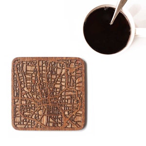 Puede incluir: Posavasos cuadrado de madera con un diseño de mapa detallado, probablemente un plano de la ciudad, en un tono marrón cálido. Una taza de café blanca con líquido oscuro y una cuchara está en el fondo. El posavasos está sobre una superficie blanca.
