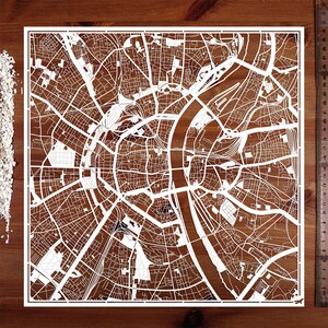 May include: A white paper cut-out of a city map, showing the streets and river of a city. The map is on a brown wooden surface.