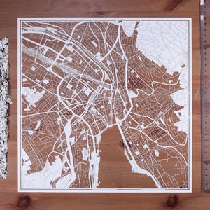 以下が含まれることがあります： 複雑な通りの詳細と建物を描いた、白い紙で切り抜かれた都市の地図。地図は木製の表面の上に置かれています。