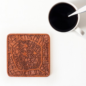 Puede incluir: Un posavasos de madera con un diseño detallado de un mapa de la ciudad cortado con láser. El posavasos es de color marrón y tiene forma cuadrada.