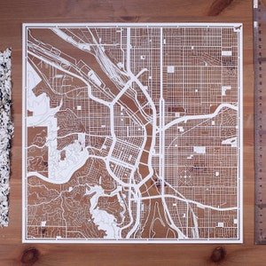 Può includere: Una mappa di una città ritagliata su carta bianca con un fiume che la attraversa. La mappa è dettagliata e mostra le strade e gli edifici della città.