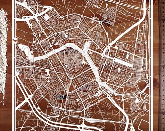 Puede incluir: Un detallado mapa blanco recortado en papel de una ciudad, sobre un fondo de madera oscura. El mapa presenta intrincados patrones de calles, vías fluviales y un borde blanco. Una regla con medidas en cm es visible.