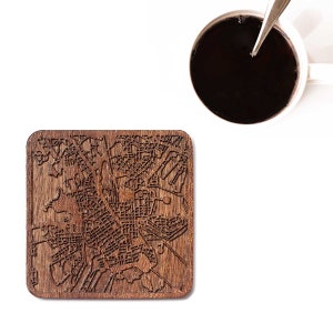 Peut inclure: Un dessous de verre carré en bois avec une carte détaillée gravée sur sa surface. Le dessous de verre est de couleur marron chaud et se trouve à côté d'une tasse blanche remplie de café noir, avec une cuillère en argent à l'intérieur. Le dessous de verre représente une carte de ville.