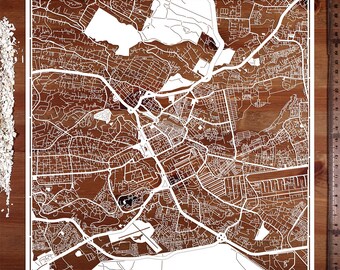 Puede incluir: Un recorte de papel blanco de un mapa de la ciudad, que muestra calles y edificios. El mapa está sobre una superficie de madera marrón.