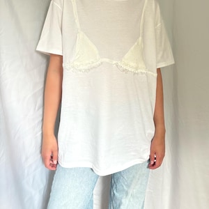 May include: A white short-sleeved t-shirt featuring a cream-colored lace bralette appliqué stitched onto the front chest area. The lace detail includes delicate scalloped edges and a textured pattern, layered over the plain white cotton fabric.