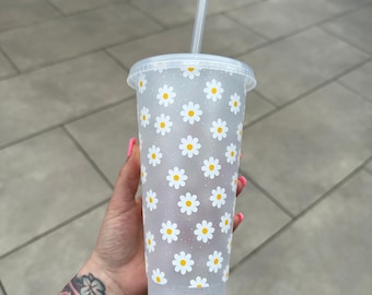 May include: A clear plastic tumbler with a white daisy pattern and a clear plastic straw. The tumbler has a glittery finish.