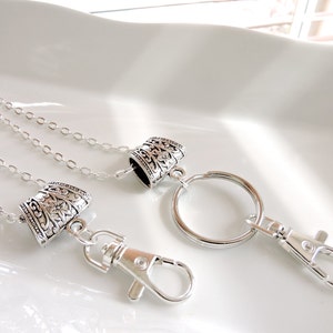 Könnte beinhalten: Silberkette mit zwei dekorativen Silberanhängern und einem silbernen Schlüsselring mit einem silbernen Karabiner.