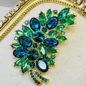 May include: A gold-tone brooch featuring a cluster of faceted teal and emerald green crystals in various shapes, including marquise, oval, and round cuts, arranged in a leaf-like pattern with two curved stems at the base set with small blue stones.