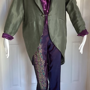 Può includere: Una lunga giacca verde in seta con collo a scialle e un unico bottone di chiusura. La giacca è indossata sopra una blusa viola e pantaloni viola con un motivo floreale.