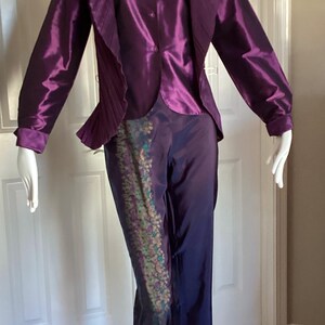May include: A purple satin pantsuit with a pleated jacket and a patterned detail on the pants.