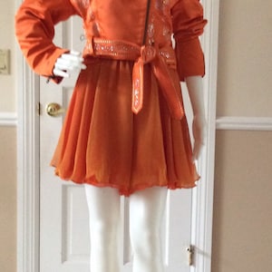 May include: A bright orange satin jacket with a zip closure and a matching orange skirt. The jacket has a decorative belt and is embellished with silver sequins.