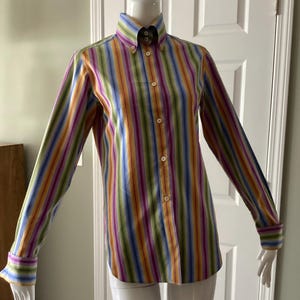 Puede incluir: Una camisa abotonada con rayas verticales en una variedad de colores, incluyendo azul, verde, amarillo, naranja y morado. La camisa de manga larga tiene cuello abotonado y puños. La camisa se exhibe en un maniquí.