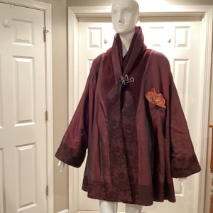 Puede incluir: Una chaqueta de color burdeos, de aspecto sedoso, con ribete negro y un cierre decorativo. La chaqueta tiene un cuello chal y un bolsillo en el lado izquierdo.