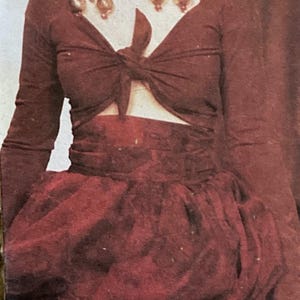 Puede incluir: Conjunto burdeos con top de manga larga anudado a la cintura y falda voluminosa. La modelo lleva pendientes grandes y ornamentados. Un conjunto de moda vintage.