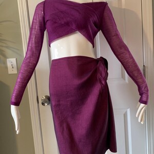 May include: A two-piece outfit in a deep purple colour. The top is a wrap-style with long sleeves, and the skirt is a knee-length wrap skirt. The fabric has a subtle sheen. The outfit is displayed on a mannequin.