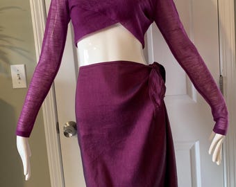 May include: A two-piece set in a deep purple hue. The top is a wrap-style with long sleeves, and the skirt is a knee-length wrap skirt. The fabric appears to have a subtle sheen. The outfit is displayed on a mannequin.
