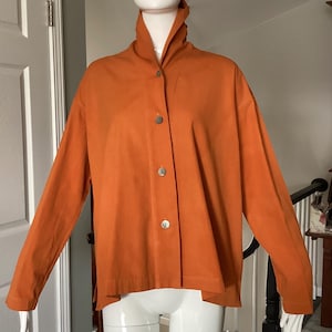 May include: An orange button-down jacket with a high collar. The jacket has long sleeves and three visible buttons down the front. The fabric appears to be a solid color, and the jacket is displayed on a mannequin.
