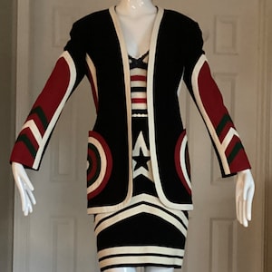 May include: A black and white striped dress with a red, white and green geometric pattern on the sleeves and skirt. The dress is being modelled by a mannequin.