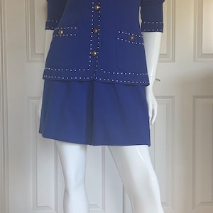 Op de afbeelding: Een koninklijk blauwe rokpak met gouden knopen en een studded trim. Het pak is op een wit mannequin.